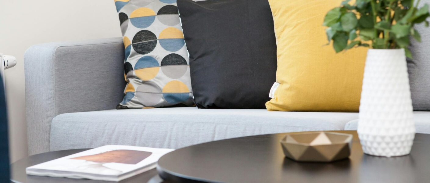A modern living room features a light grey sofa with three cushions—one patterned with geometric shapes in yellow, grey, and black; one solid black; and one solid yellow. In front, two black tables hold a white vase of white roses, a small golden bowl, and a magazine.