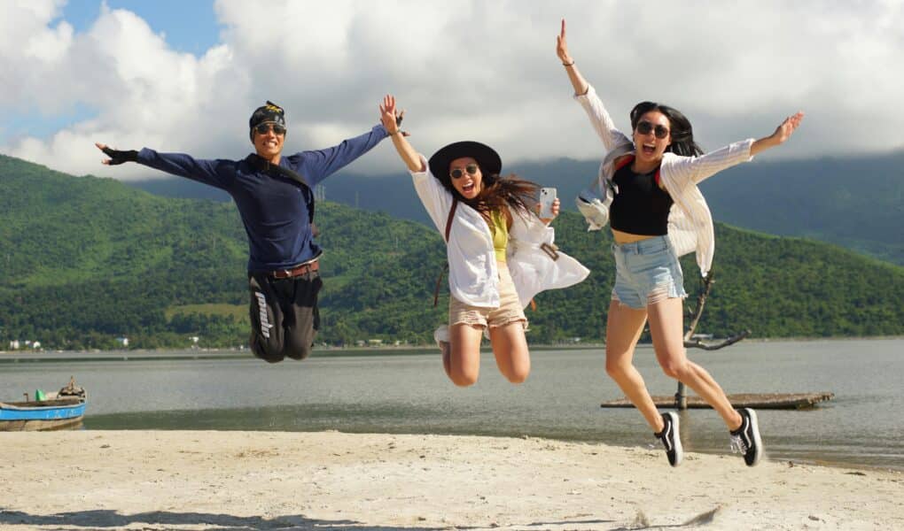 Three people are joyfully jumping in the air on a sandy beach, arms raised. Behind them are green hills, a calm body of water, and a partly cloudy sky. All three wear sunglasses and casual summer clothes, smiling and looking happy.