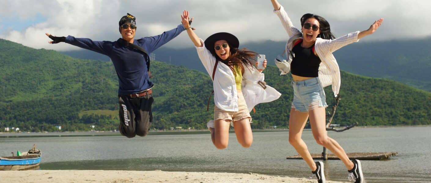 Three people are joyfully jumping in the air on a sandy beach, arms raised. Behind them are green hills, a calm body of water, and a partly cloudy sky. All three wear sunglasses and casual summer clothes, smiling and looking happy.