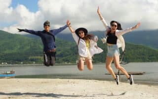 Three people are joyfully jumping in the air on a sandy beach, arms raised. Behind them are green hills, a calm body of water, and a partly cloudy sky. All three wear sunglasses and casual summer clothes, smiling and looking happy.
