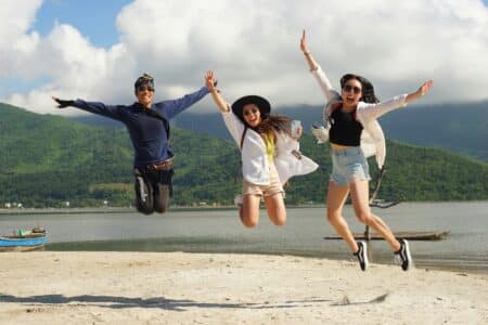 Three people are joyfully jumping in the air on a sandy beach, arms raised. Behind them are green hills, a calm body of water, and a partly cloudy sky. All three wear sunglasses and casual summer clothes, smiling and looking happy.