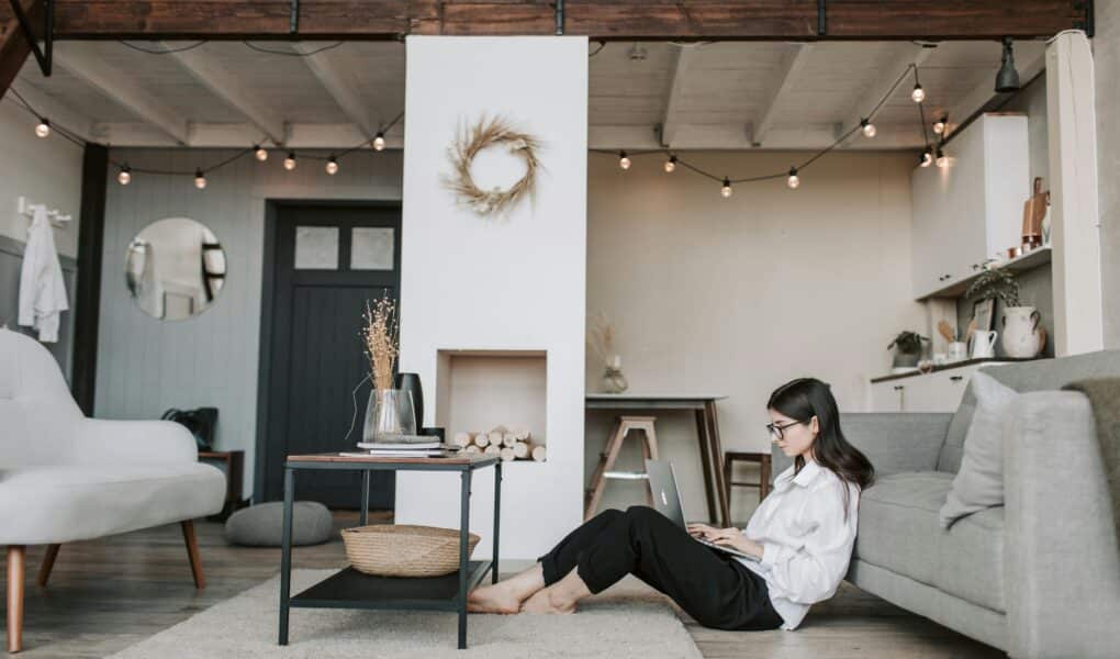 A woman with long dark hair, wearing glasses and a white shirt, sits on the floor leaning against a grey sofa, working on a laptop. The cosy, modern flat features fairy lights, a loft bed, minimalist furniture, and neutral colours.