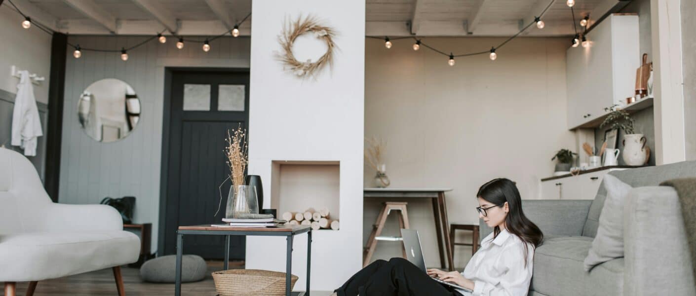 A woman with long dark hair, wearing glasses and a white shirt, sits on the floor leaning against a grey sofa, working on a laptop. The cosy, modern flat features fairy lights, a loft bed, minimalist furniture, and neutral colours.