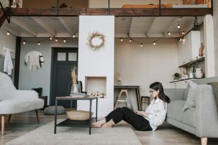 A woman with long dark hair, wearing glasses and a white shirt, sits on the floor leaning against a grey sofa, working on a laptop. The cosy, modern flat features fairy lights, a loft bed, minimalist furniture, and neutral colours.