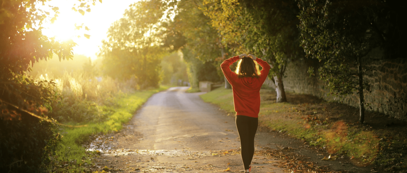 A person in a red jumper and black trousers walks alone down a sunlit, tree-lined path covered with fallen leaves, holding their hands behind their head. The sun is low, casting a warm golden light through the trees on the left.