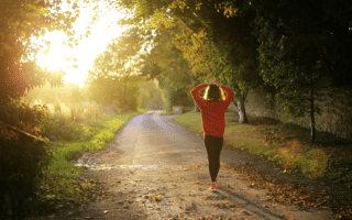 A person in a red jumper and black trousers walks alone down a sunlit, tree-lined path covered with fallen leaves, holding their hands behind their head. The sun is low, casting a warm golden light through the trees on the left.