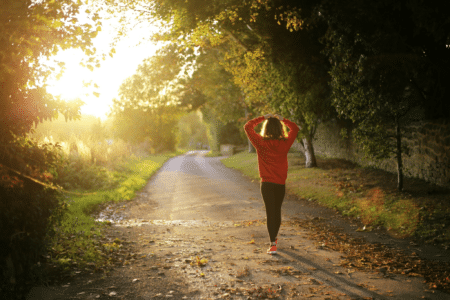 A person in a red jumper and black trousers walks alone down a sunlit, tree-lined path covered with fallen leaves, holding their hands behind their head. The sun is low, casting a warm golden light through the trees on the left.