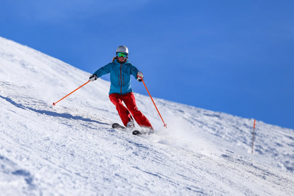 A skier in a blue jacket, red trousers, helmet, and goggles glides downhill on a snowy slope, holding orange ski poles. The sky is clear and blue, and snow sprays up from the skier’s movement. A slope marker is visible in the background.