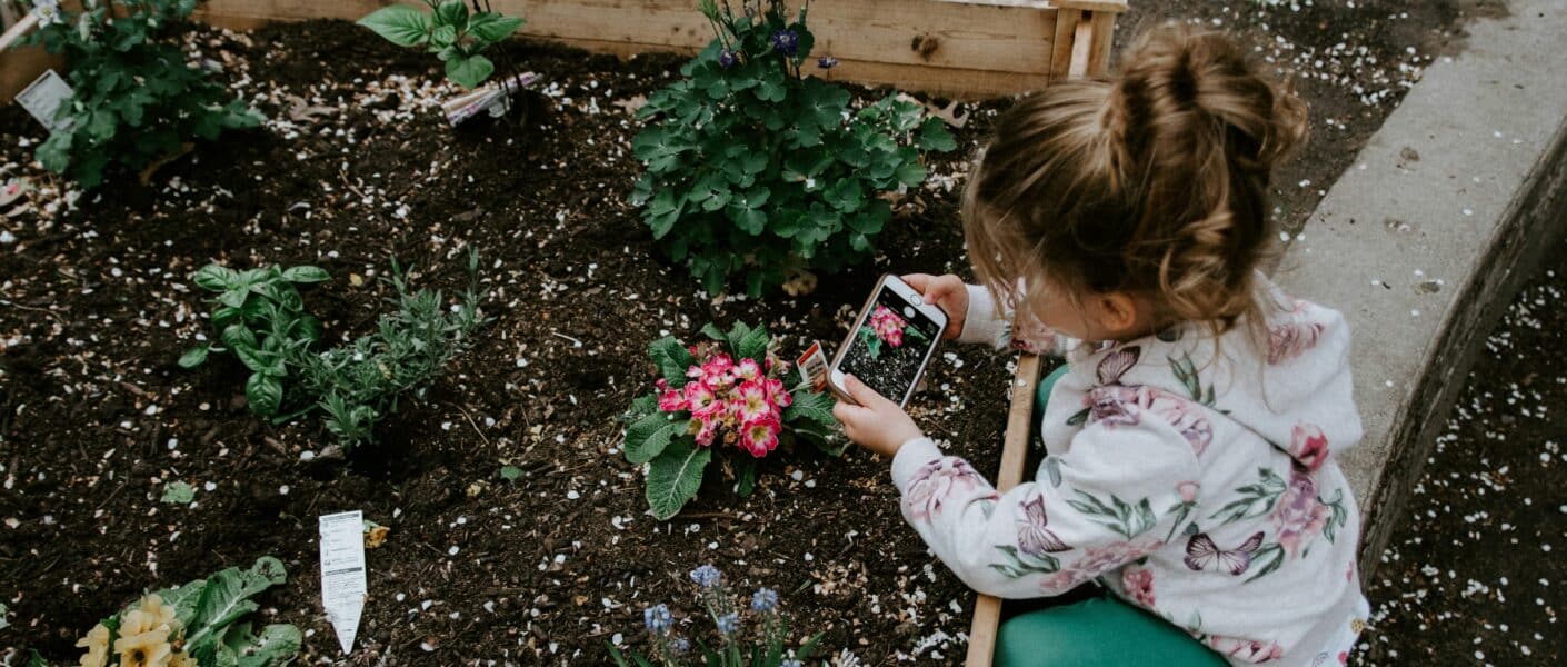 A young girl wearing a floral hoodie and teal trousers kneels by a raised garden bed, taking a photo of pink flowers with a mobile phone. Around her are small blooming plants and garden markers in the freshly turned soil.