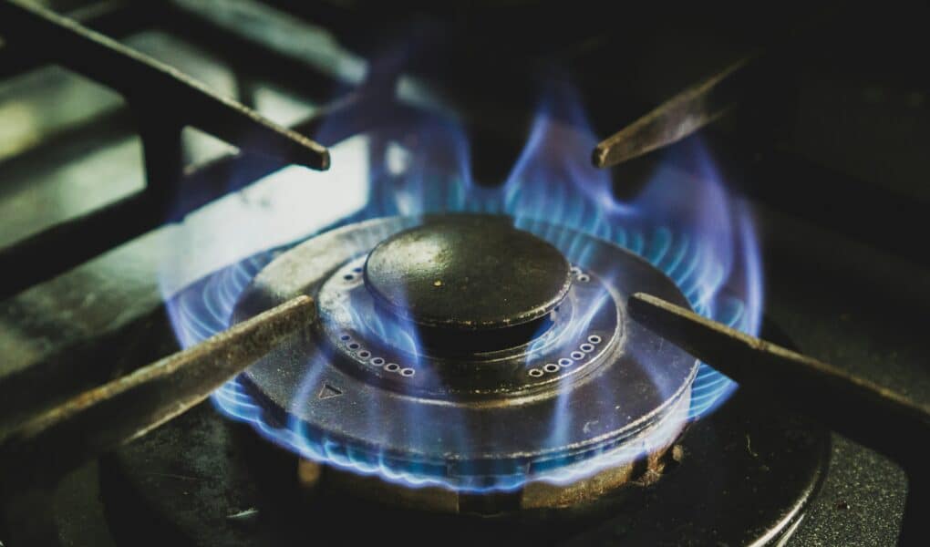 Close-up of a lit gas hob burner with blue flames evenly circling the burner cap. The metal pan supports of the hob are visible around the burner. The dark, slightly worn burner contrasts with the glowing blue flames, indicating it is actively in use.