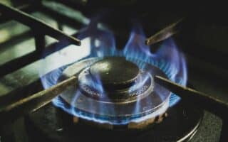 Close-up of a lit gas hob burner with blue flames evenly circling the burner cap. The metal pan supports of the hob are visible around the burner. The dark, slightly worn burner contrasts with the glowing blue flames, indicating it is actively in use.