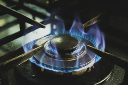 Close-up of a lit gas hob burner with blue flames evenly circling the burner cap. The metal pan supports of the hob are visible around the burner. The dark, slightly worn burner contrasts with the glowing blue flames, indicating it is actively in use.