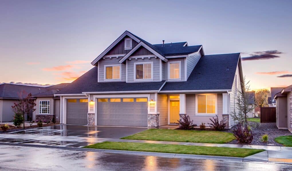 A modern two-storey house with grey cladding, white trim, and a three-car garage. Warm lights glow from the windows. The driveway and street are wet, reflecting the house, suggesting recent rain. The sky is colourful with clouds at sunset.