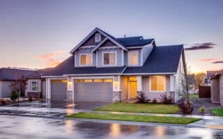 A modern two-storey house with grey cladding, white trim, and a three-car garage. Warm lights glow from the windows. The driveway and street are wet, reflecting the house, suggesting recent rain. The sky is colourful with clouds at sunset.