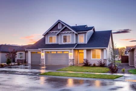 A modern two-storey house with grey cladding, white trim, and a three-car garage. Warm lights glow from the windows. The driveway and street are wet, reflecting the house, suggesting recent rain. The sky is colourful with clouds at sunset.