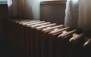 A close-up view of a vintage, brown metal radiator positioned in front of a window with sheer white curtains gently illuminated by soft, natural light filtering through the glass. The radiator’s surface shows subtle wear and texture.