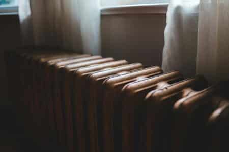 A close-up view of a vintage, brown metal radiator positioned in front of a window with sheer white curtains gently illuminated by soft, natural light filtering through the glass. The radiator’s surface shows subtle wear and texture.