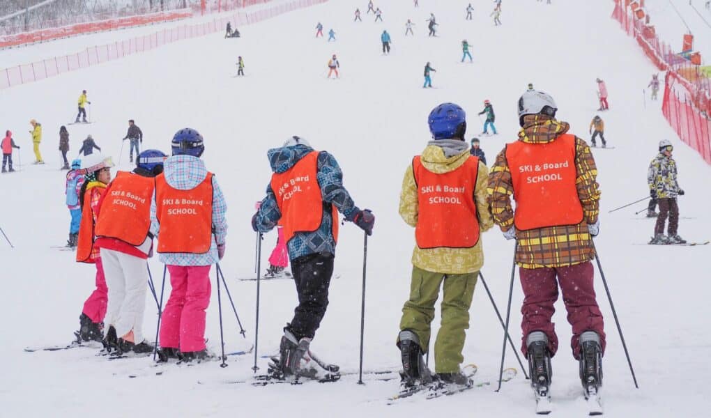 A group of children in bright orange vests labelled "SKI & BOARD SCHOOL" stand with skis on a snowy slope, facing away. More people ski in the background on the wide, snow-covered hill bordered by red fencing, with trees visible in the distance.