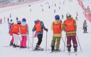 A group of children in bright orange vests labelled "SKI & BOARD SCHOOL" stand with skis on a snowy slope, facing away. More people ski in the background on the wide, snow-covered hill bordered by red fencing, with trees visible in the distance.