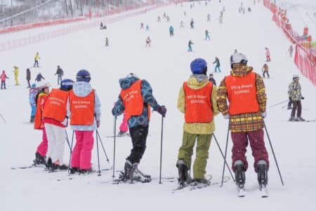 A group of children in bright orange vests labelled "SKI & BOARD SCHOOL" stand with skis on a snowy slope, facing away. More people ski in the background on the wide, snow-covered hill bordered by red fencing, with trees visible in the distance.