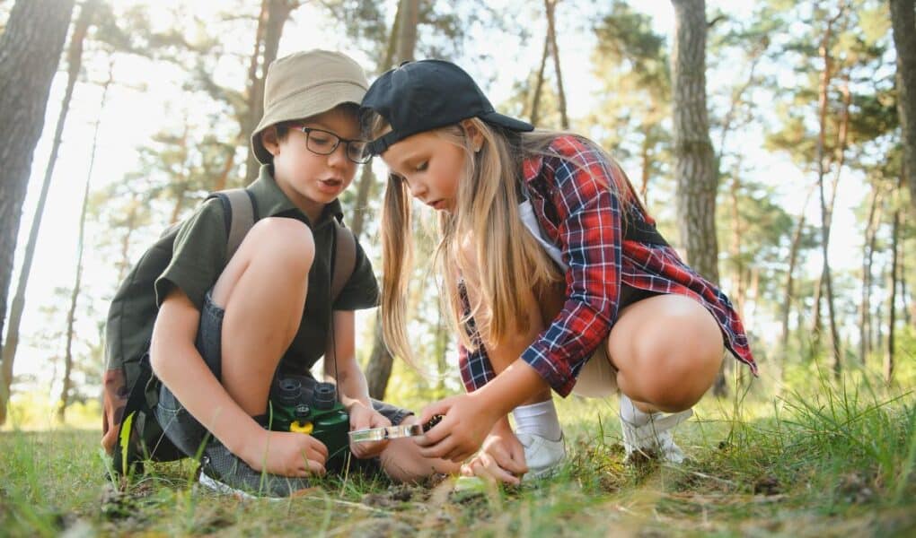 Two children in a forest examine something on the ground with a magnifying glass. The boy wears glasses, a hat, and a rucksack with binoculars, while the girl wears a black cap and red checked shirt. Sunlight filters through tall trees around them.