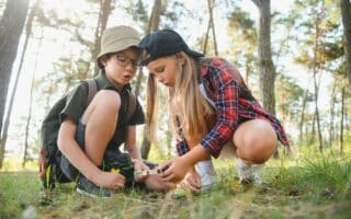 Two children in a forest examine something on the ground with a magnifying glass. The boy wears glasses, a hat, and a rucksack with binoculars, while the girl wears a black cap and red checked shirt. Sunlight filters through tall trees around them.