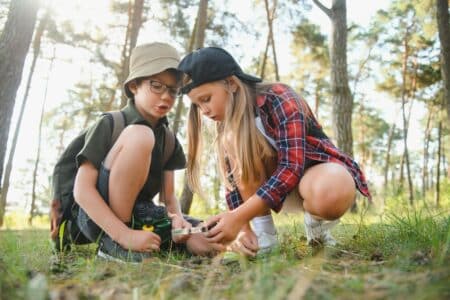 Two children in a forest examine something on the ground with a magnifying glass. The boy wears glasses, a hat, and a rucksack with binoculars, while the girl wears a black cap and red checked shirt. Sunlight filters through tall trees around them.