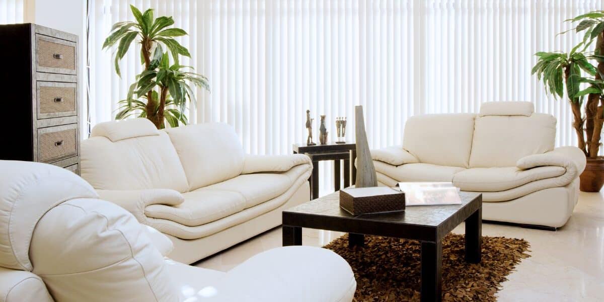 Modern living room with three cream-coloured leather sofas around a dark rectangular coffee table on a brown textured rug. Tall potted plants, decorative vases on the table, and vertical blinds cover large windows, filling the room with natural light.