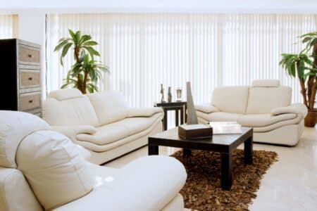 Modern living room with three cream-coloured leather sofas around a dark rectangular coffee table on a brown textured rug. Tall potted plants, decorative vases on the table, and vertical blinds cover large windows, filling the room with natural light.