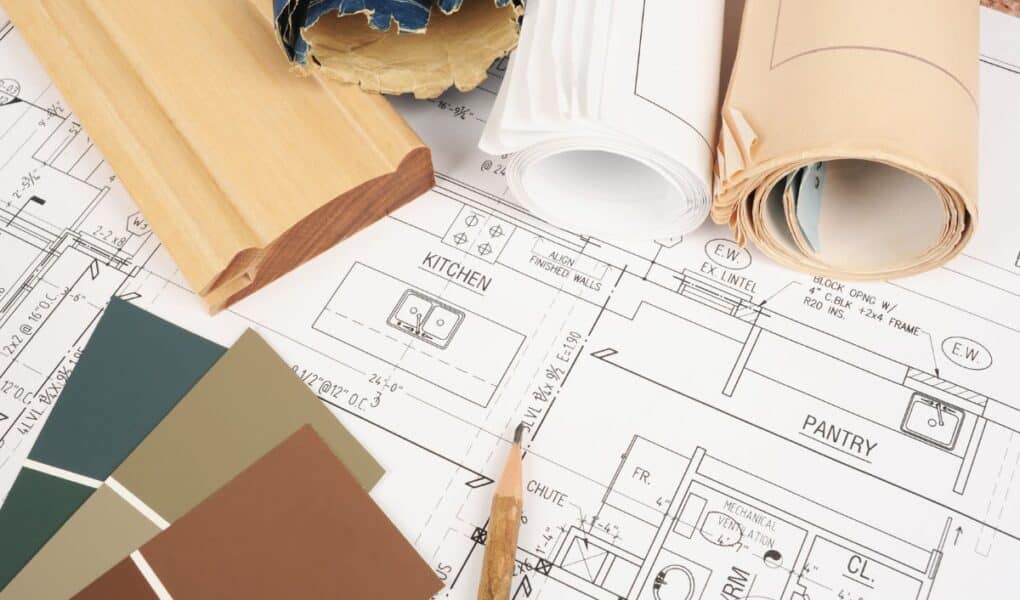 A close-up of architectural blueprints showing kitchen and larder layouts, with rolled-up plans, a wooden trim sample, and green paint swatches arranged on top, suggesting interior design or home renovation planning.