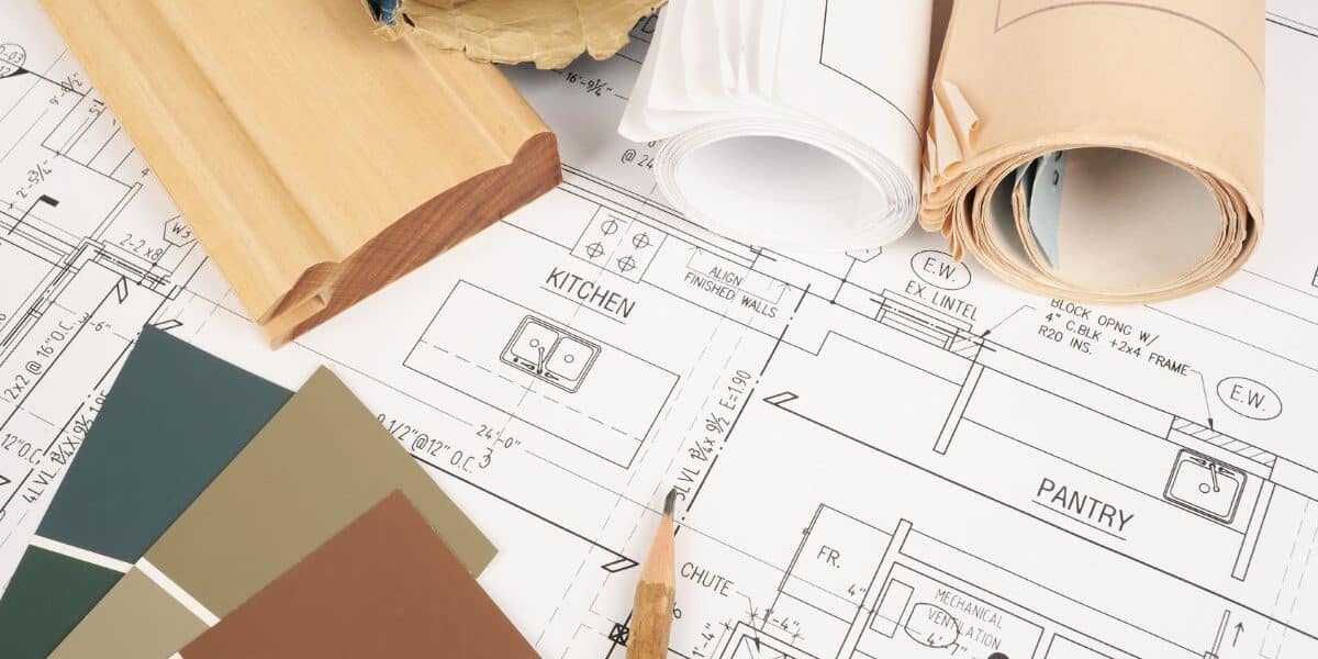 A close-up of architectural blueprints showing kitchen and larder layouts, with rolled-up plans, a wooden trim sample, and green paint swatches arranged on top, suggesting interior design or home renovation planning.