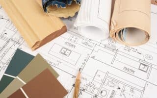 A close-up of architectural blueprints showing kitchen and larder layouts, with rolled-up plans, a wooden trim sample, and green paint swatches arranged on top, suggesting interior design or home renovation planning.