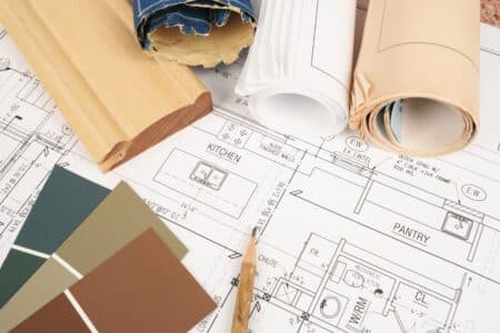 A close-up of architectural blueprints showing kitchen and larder layouts, with rolled-up plans, a wooden trim sample, and green paint swatches arranged on top, suggesting interior design or home renovation planning.