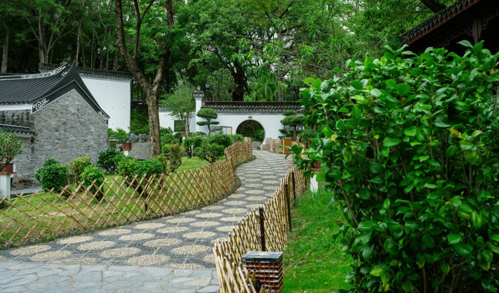 A winding stone path bordered by bamboo fences leads through a lush green Chinese garden with manicured bushes, traditional white walls, a tiled roof, and a round moon gate under tall trees. The scene is serene and well-maintained.
