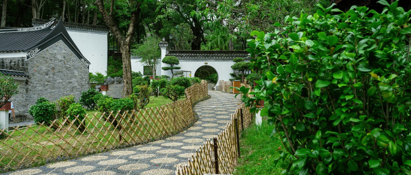 A winding stone path bordered by bamboo fences leads through a lush green Chinese garden with manicured bushes, traditional white walls, a tiled roof, and a round moon gate under tall trees. The scene is serene and well-maintained.