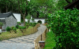A winding stone path bordered by bamboo fences leads through a lush green Chinese garden with manicured bushes, traditional white walls, a tiled roof, and a round moon gate under tall trees. The scene is serene and well-maintained.