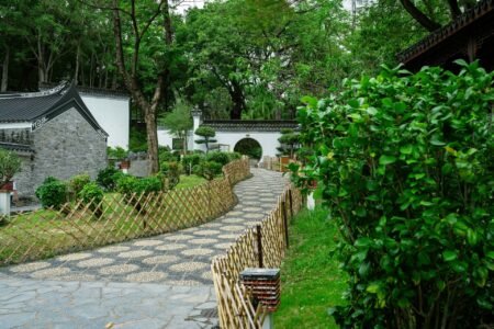 A winding stone path bordered by bamboo fences leads through a lush green Chinese garden with manicured bushes, traditional white walls, a tiled roof, and a round moon gate under tall trees. The scene is serene and well-maintained.