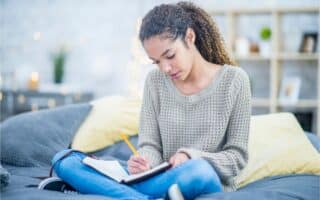 A young woman with curly hair sits cross-legged on a bed, wearing a beige jumper and jeans. She writes in a notebook with a pencil, focused on her work. The room is softly lit with decorative lights and shelves in the background.