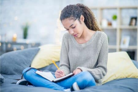 A young woman with curly hair sits cross-legged on a bed, wearing a beige jumper and jeans. She writes in a notebook with a pencil, focused on her work. The room is softly lit with decorative lights and shelves in the background.