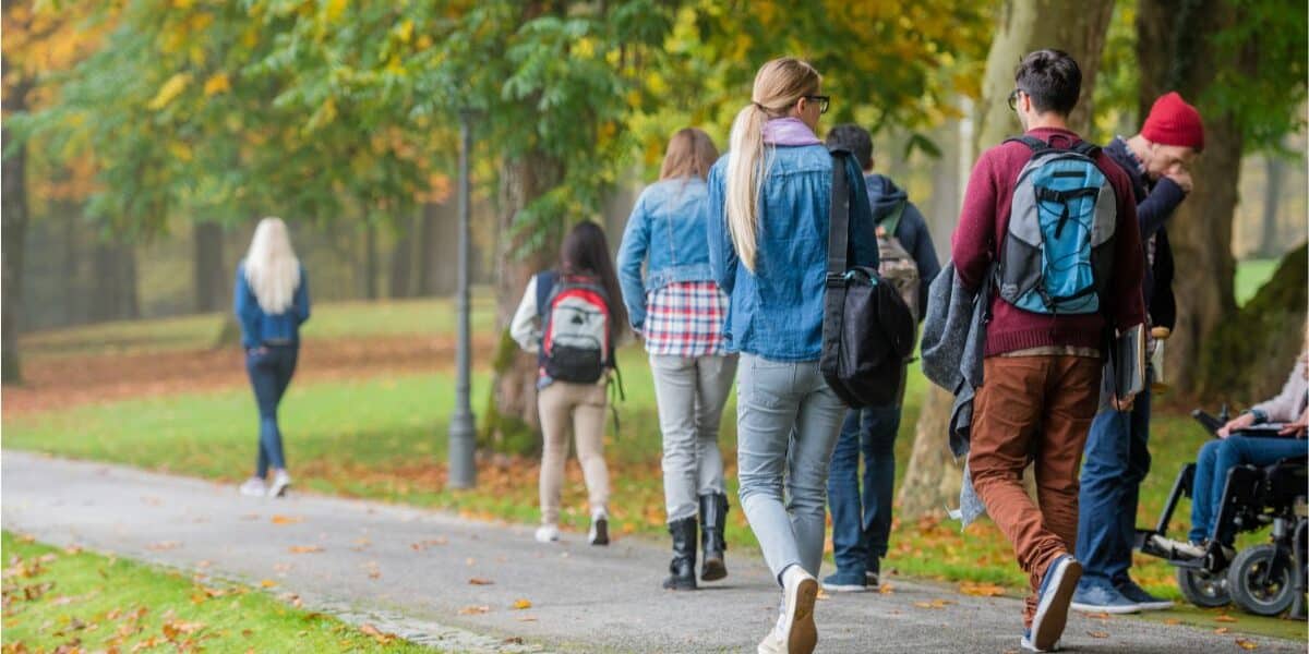 A group of people, mostly young adults, walk on a tarmac path through a park with green grass and autumn trees. Some wear rucksacks and jackets. One person uses a wheelchair. Fallen leaves are scattered on the ground.