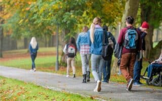 A group of people, mostly young adults, walk on a tarmac path through a park with green grass and autumn trees. Some wear rucksacks and jackets. One person uses a wheelchair. Fallen leaves are scattered on the ground.