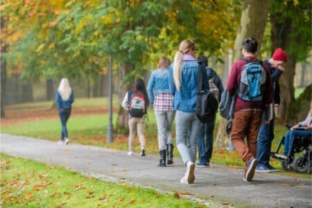 A group of people, mostly young adults, walk on a tarmac path through a park with green grass and autumn trees. Some wear rucksacks and jackets. One person uses a wheelchair. Fallen leaves are scattered on the ground.