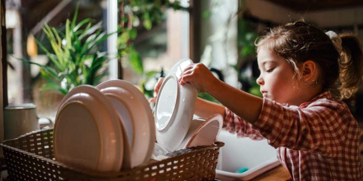A young child in a checked shirt places clean plates into a draining rack by a sunlit kitchen window, surrounded by green plants. The scene feels warm and cosy, with natural light highlighting the crockery and the child’s focused expression.