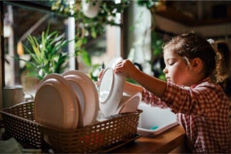 A young child in a checked shirt places clean plates into a draining rack by a sunlit kitchen window, surrounded by green plants. The scene feels warm and cosy, with natural light highlighting the crockery and the child’s focused expression.
