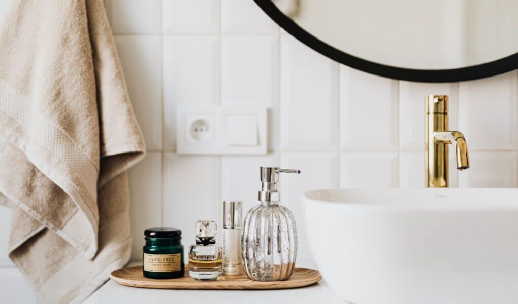 A modern bathroom worktop with a gold tap, a white basin, and a round mirror. On the worktop is a wooden tray holding a glass soap dispenser, perfume bottles, and a green jar. Beige towels hang on the wall with white tiled splashback.