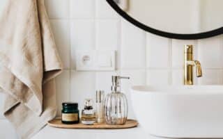 A modern bathroom worktop with a gold tap, a white basin, and a round mirror. On the worktop is a wooden tray holding a glass soap dispenser, perfume bottles, and a green jar. Beige towels hang on the wall with white tiled splashback.