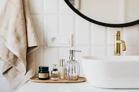 A modern bathroom worktop with a gold tap, a white basin, and a round mirror. On the worktop is a wooden tray holding a glass soap dispenser, perfume bottles, and a green jar. Beige towels hang on the wall with white tiled splashback.
