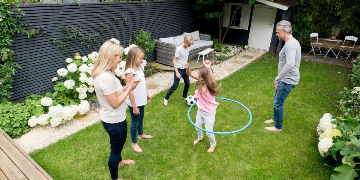 Five people, including two adults and three children, are playing in a green back garden. One child is spinning a blue hula hoop whilst others watch or play near a black-and-white football. There are white flowers, a garden shed, and outdoor seating in the garden.