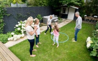 Five people, including two adults and three children, are playing in a green back garden. One child is spinning a blue hula hoop whilst others watch or play near a black-and-white football. There are white flowers, a garden shed, and outdoor seating in the garden.