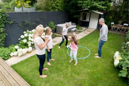 Five people, including two adults and three children, are playing in a green back garden. One child is spinning a blue hula hoop whilst others watch or play near a black-and-white football. There are white flowers, a garden shed, and outdoor seating in the garden.